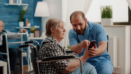 Man nurse teaching retired woman with disability to use smartphone for communication in nursing...