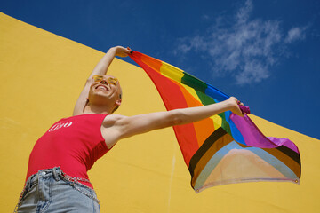Low angle view of a young non-binary person holding and raising up a rainbow flag outdoors.