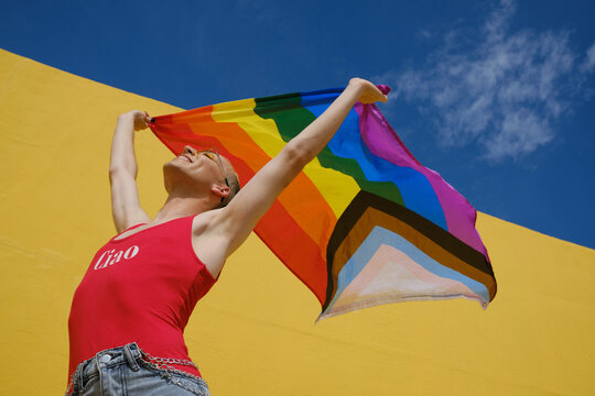 Cheerful non-binary person holding up and waving an lgbt pride flag outdoors.