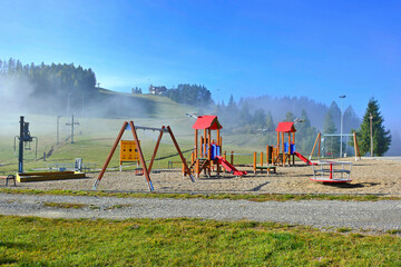 Fototapeta premium Vast green grassland and children's play area,let visitor take a deep breath in the embrace of nature. Foggy mountains on the background, Czorsztyn-Ski, Poland