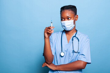 African american specialist nurse with protective face mask against covid19 holding syringe analyzing medical solution working at healthcare treatment in studio with blue background. Medicine concept