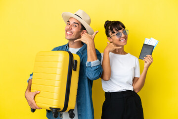 Young traveler friends holding a suitcase and passport isolated on yellow background making phone gesture. Call me back sign
