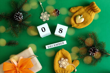 Calendar for December 8 : the name of the month in English, cubes with numbers 0 and 8, yellow mittens, a gift with a yellow bow, spruce branches on a green background, top view, bokeh