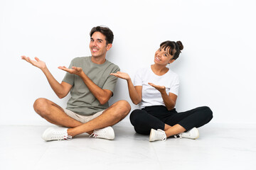 Fototapeta na wymiar Young mixed race couple sitting on the floor isolated on white background inviting to come with hand. Happy that you came