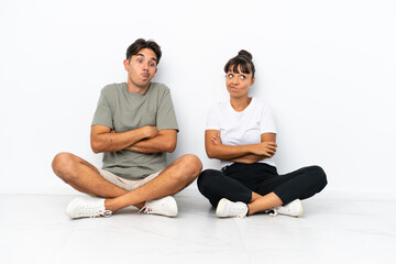 Fototapeta na wymiar Young mixed race couple sitting on the floor isolated on white background making doubts gesture while lifting the shoulders