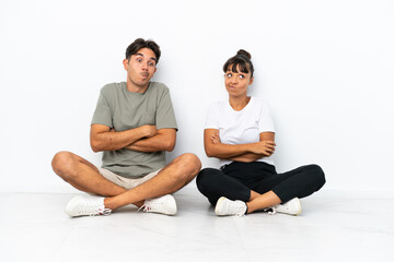 Young mixed race couple sitting on the floor isolated on white background making doubts gesture while lifting the shoulders