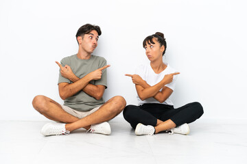 Fototapeta premium Young mixed race couple sitting on the floor isolated on white background pointing to the laterals having doubts