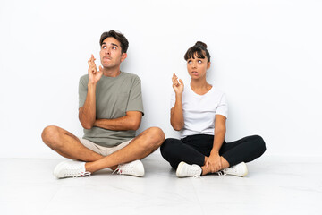 Fototapeta na wymiar Young mixed race couple sitting on the floor isolated on white background with fingers crossing and wishing the best