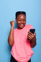 Portrait of happy african american young woman received a message from friend enjoying good news standing in studio with blue background. Teenager using smartphone browsing on social media