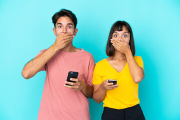 Young mixed race couple holding mobile phone isolated on blue background covering mouth with hands for saying something inappropriate - obrazy, fototapety, plakaty