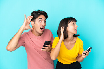 Young mixed race couple holding mobile phone isolated on blue background listening to something by putting hand on the ear