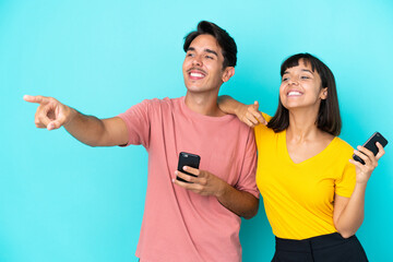 Young mixed race couple holding mobile phone isolated on blue background presenting an idea while looking smiling towards