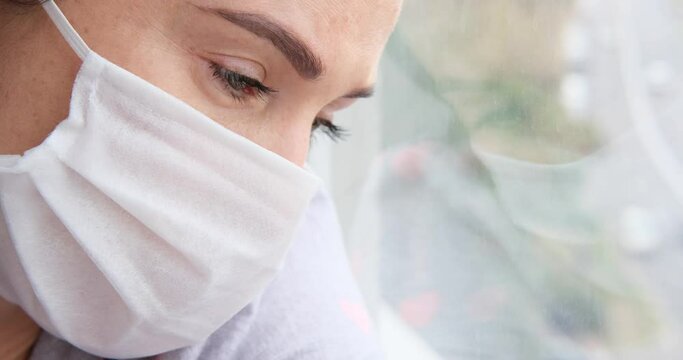 Sad self isolated woman wearing protective face mask leaning against window while recovering at home