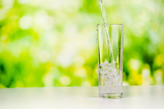 Pouring Pure Water Into A Tall Transparent Glass On The Desk With A Greenery Background. A Studio Shoot Of Water Pouring.