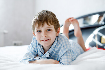 Adorable happy little kid boy after sleeping in his white bed in colorful nightwear. School child celebrating pajama party and looking at the camera. Funny happy child playing and smiling