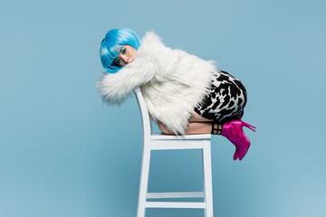 Stylish asian woman in wig, heels and fluffy jacket posing on chair isolated on blue
