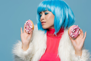 Stylish asian woman in bright wig holding delicious donuts isolated on blue
