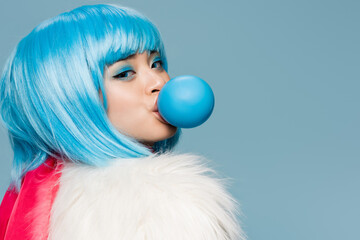 Pretty asian woman in furry jacket blowing bubble gum and looking at camera isolated on blue