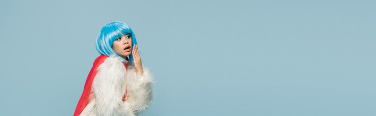 Astonished asian woman in wig and furry jacket looking away isolated on blue, banner