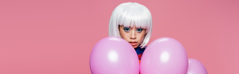 Asian woman in white wig looking at camera near balloons isolated on pink, banner