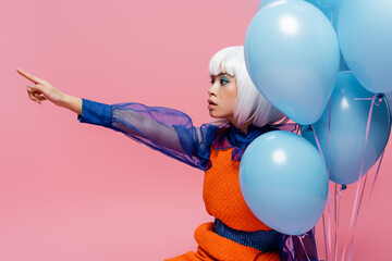 Asian pop art woman pointing with finger near blue balloons isolated on pink