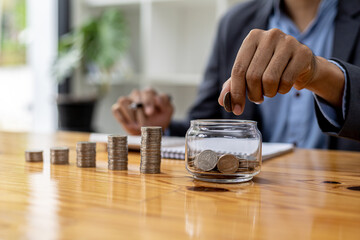 Fototapeta na wymiar A man is dropping coins into a glass jar with a number of coins inside, on a desk are stacked coins from low to high showing the growth of money. Concept of saving and investing money.