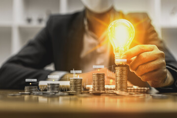 A businessman holds a glowing light bulb on top of the highest pile of coins. Placing coins in a row from low to high is comparable to saving money to grow more. Savings growth concept. © kamiphotos