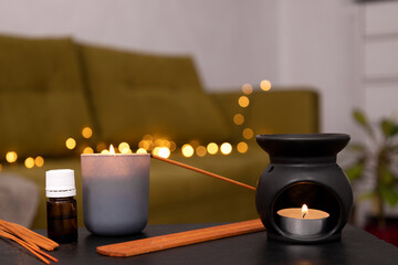 Aromatic essential oil bottle,candle,aroma lamp and incense sticks on the dark table against home sofa,plant and cozy warm light.Empty space