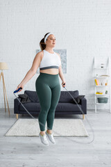 Young plus size woman jumping with skipping rope in living room - obrazy, fototapety, plakaty