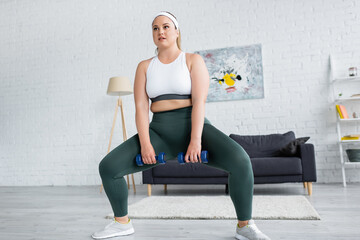 Plus size woman doing squat while training with dumbbells at home