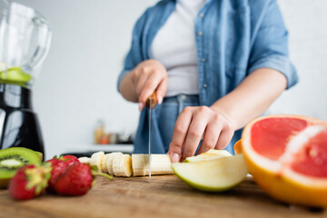 Cropped view of blurred woman with overweight cutting banana near fruits and blender in kitchen