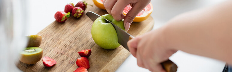 Cropped view of young woman cutting apple near fruits on cutting board in kitchen, banner