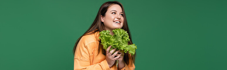 Cheerful woman with overweight holding organic lettuce isolated on green, banner