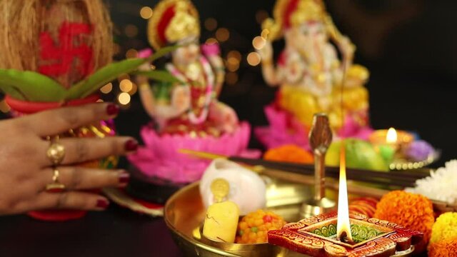 Square Clay Diya Deep Or Dia Lit In Puja Thali With Ganesha Lakshmi Laxmi In Background. Hands Of Girl Wearing Choodi Places Kalash And Fruits. Theme For Dussehra Puja, Shubh Deepawali