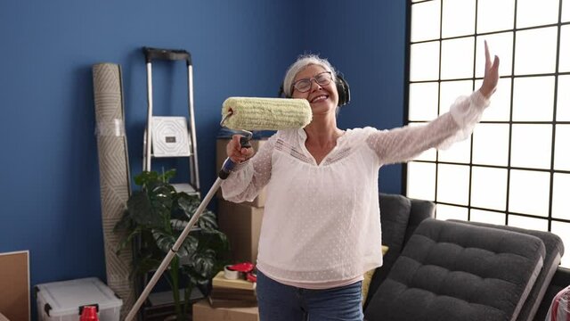Middle age grey-haired woman singing song using paint roller as a microphone at new home
