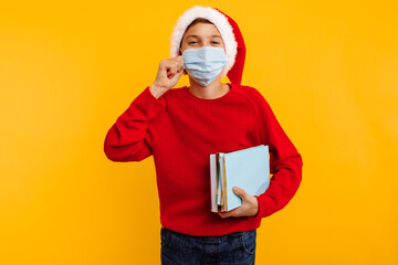 boy in a protective medical mask with books and a hat of santa claus, on a yellow background. online education
