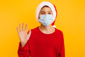 boy in protective medical mask and santa claus hat, waving hello to the camera, on a yellow background. communicates