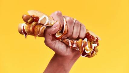 Female hand squeezing junk hot dog with mustard and ketchup isolated over yellow background