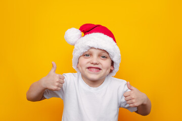 A little cute caucasian cheerful kid in a white t-shirt and a red Santa's hat shows thumb up like gesture with his hands isolated on a color yellow background. Christmas and New Year concept