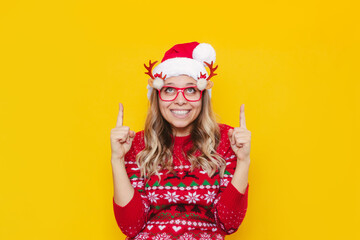 A young smiling blonde woman in glasses with deer antlers, a red deer sweater and Christmas Santa hat points up to empty copy space for greeting text with fingers isolated on color yellow background