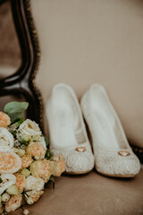 Wedding shoes of the bride with a bouquet of flowers on a beige armchair with wedding rings.
