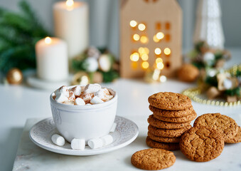 Hot cocoa with marshmallow in white mug surrounded by winter Christmas decoration on white table. Cosy holidays and New Year concept. Selective focus, blurred kraft paper house with garland