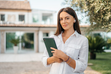 Fototapeta premium Portrait of a happy brunette woman smiling holding a tablet in the hands of a summer garden white shirt. The realtor presents and sells the house