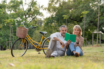 Happy couple senior sit on grass relaxing and reading guest book together after riding bicycle in...