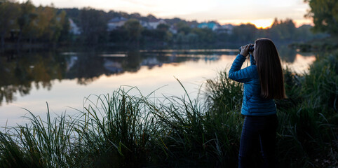 Young birdwatcher woman standing with binoculars near the pond in the evening
