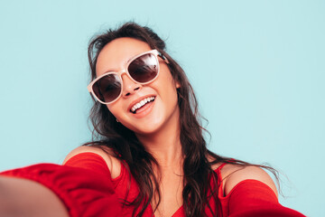 Young beautiful smiling female in trendy summer red top and jeans clothes. Sexy carefree woman posing near blue wall in studio. Positive brunette model having fun. Taking Pov selfie in sunglasses