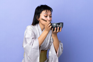 Young woman wearing kimono over isolated blue background holding a bowl of noodles with chopsticks and eating it