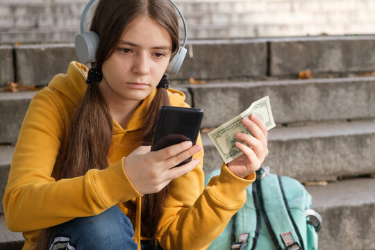 The teenager holds dollar bills and looks at the smartphone screen.