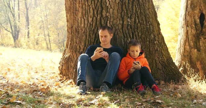 Father And Son Rests Under the Huge Oak in National Park While Hiking in Autumn. Family Use Smartphones in Nature and Talk with Each Other