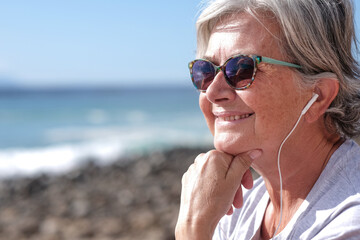 Portrait of a senior smiling woman sitting on the beach. Listening music with earphones - horizon over sea